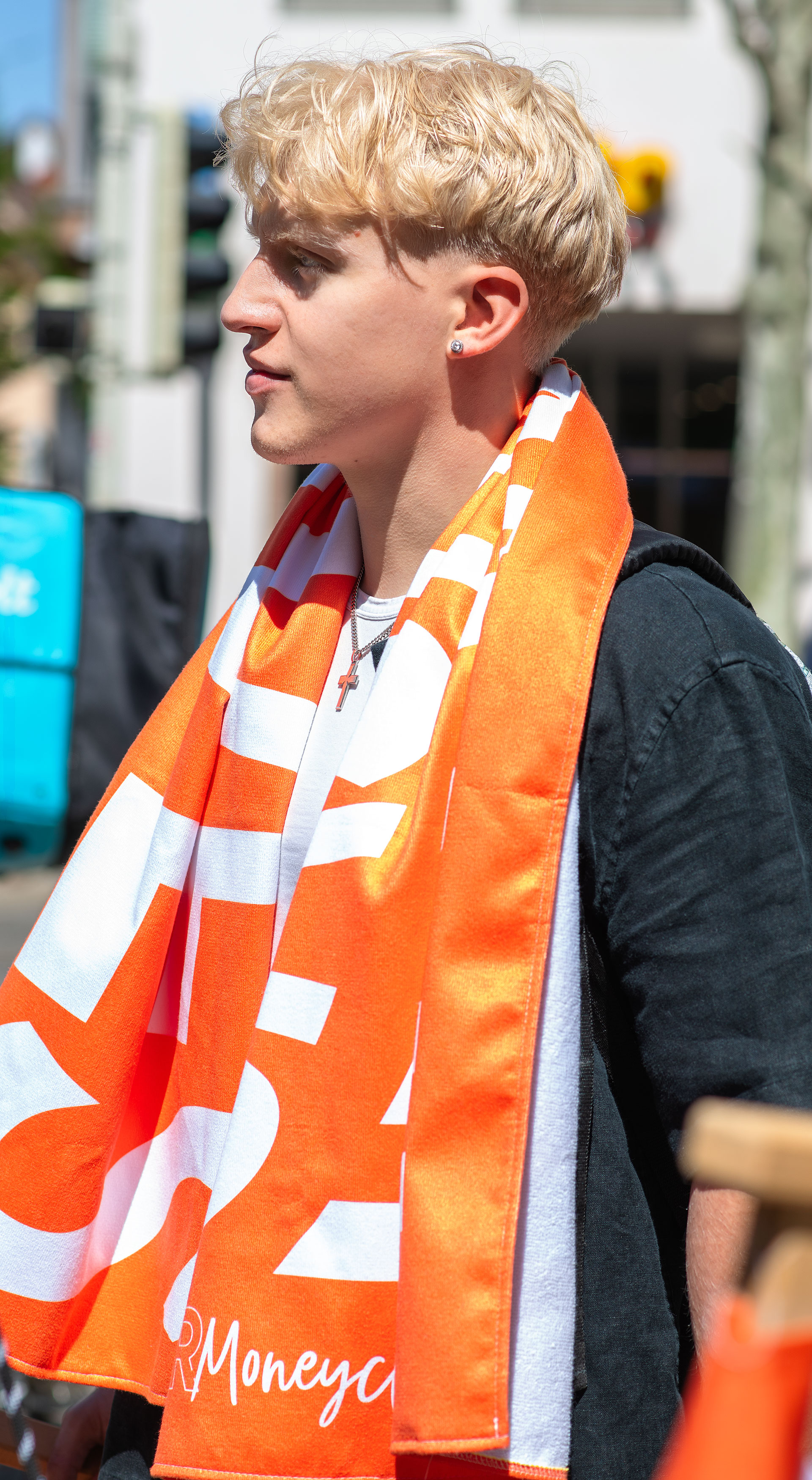 Seitenprofil einer Person mit blonden Haaren, die ein orange-weißes Handtuch mit dem „VR Moneyclub“-Schriftzug locker um den Nacken trägt, aufgenommen in einem urbanen Außenbereich.Seitenprofil einer Person mit blonden Haaren, die ein orange-weißes Handtuch mit dem „VR Moneyclub“-Schriftzug locker um den Nacken trägt, aufgenommen in einem urbanen Außenbereich.