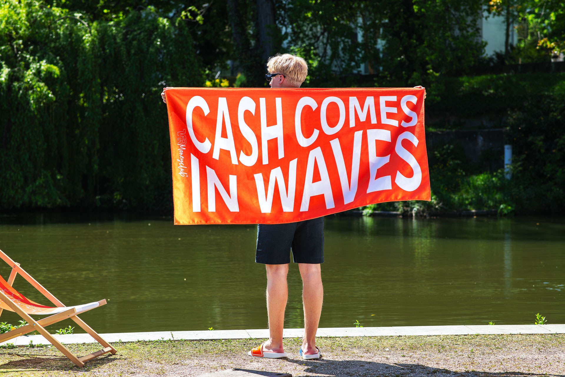 Rückansicht einer Person, die an einem sonnigen Ufer steht und ein großes orangefarbenes Handtuch mit dem auffälligen weißen Schriftzug „CASH COMES IN WAVES“ und dem dezenten „VR Moneyclub“-Logo hochhält.