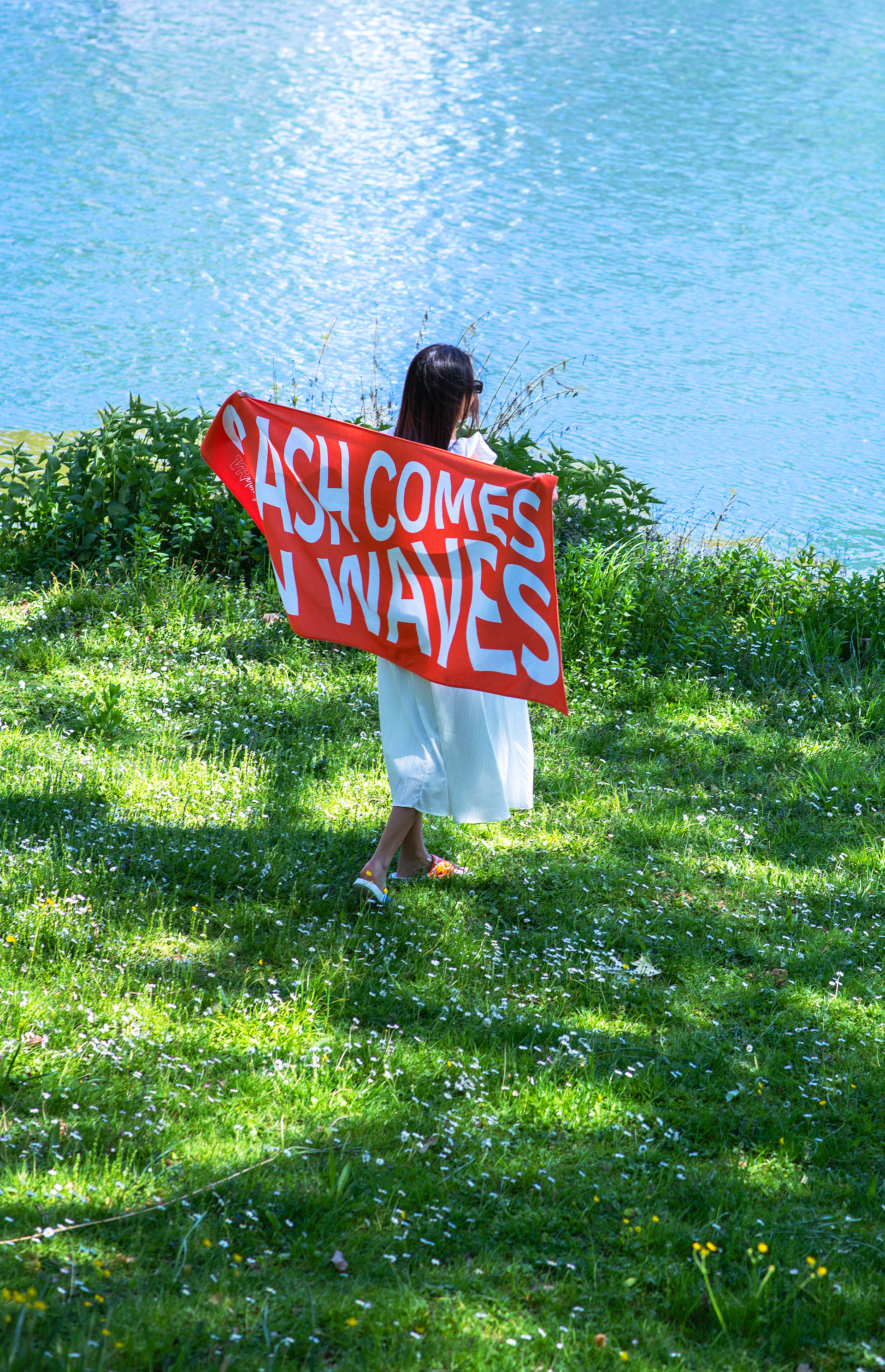 Rückansicht einer Person, die auf einer blühenden Wiese an einem Gewässer steht und ein großes, leuchtend orangefarbenes Handtuch mit dem weißen Slogan „CASH COMES IN WAVES“ und dem „VR Moneyclub“-Logo präsentiert.