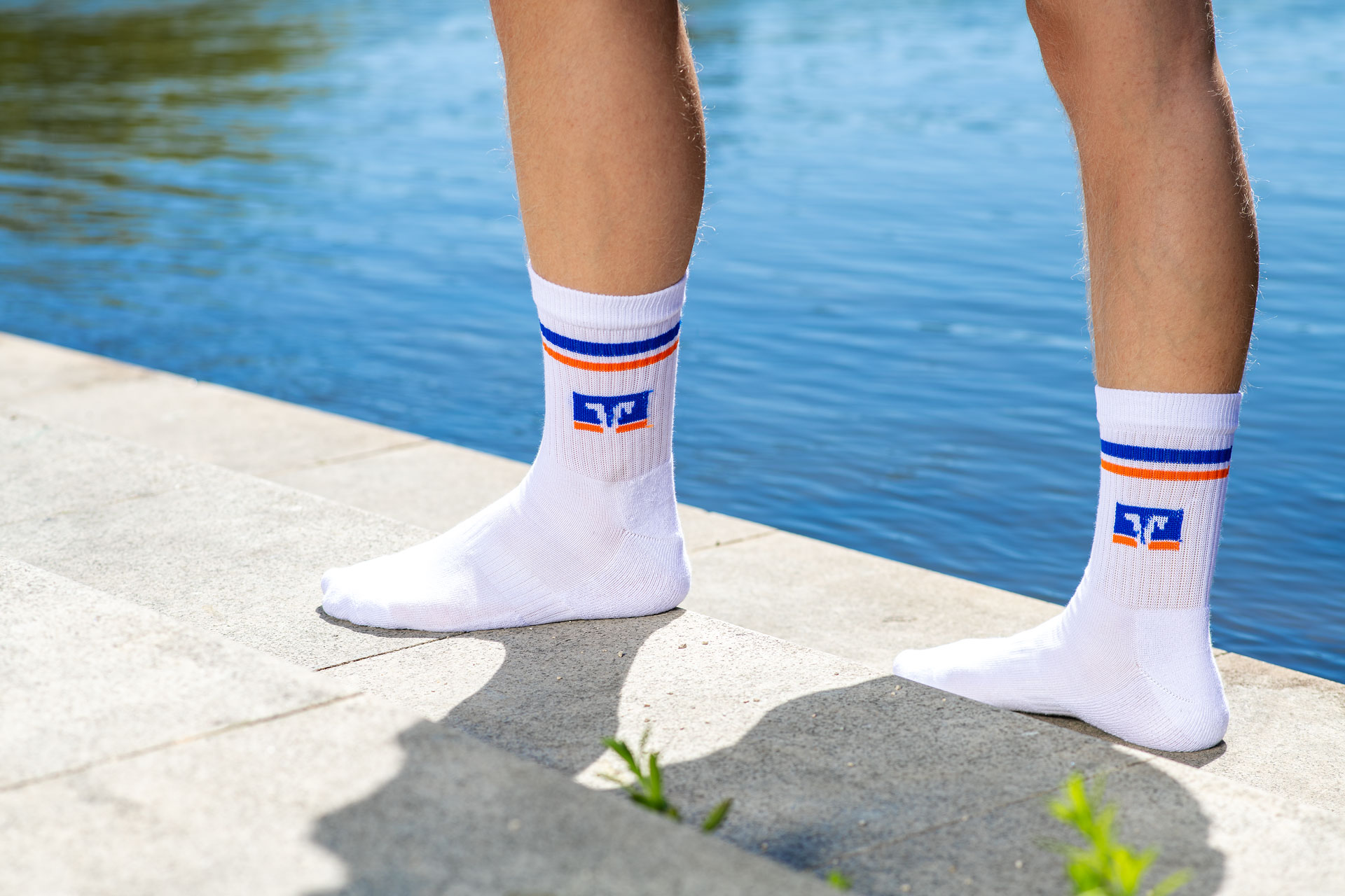 Nahaufnahme der Füße und Unterschenkel einer Person, die weiße Crew-Socken mit blau-orangen Streifen und einem VR-Logo trägt, stehend auf einer Steinstufe neben blauem Wasser in einer sonnigen Außenumgebung.