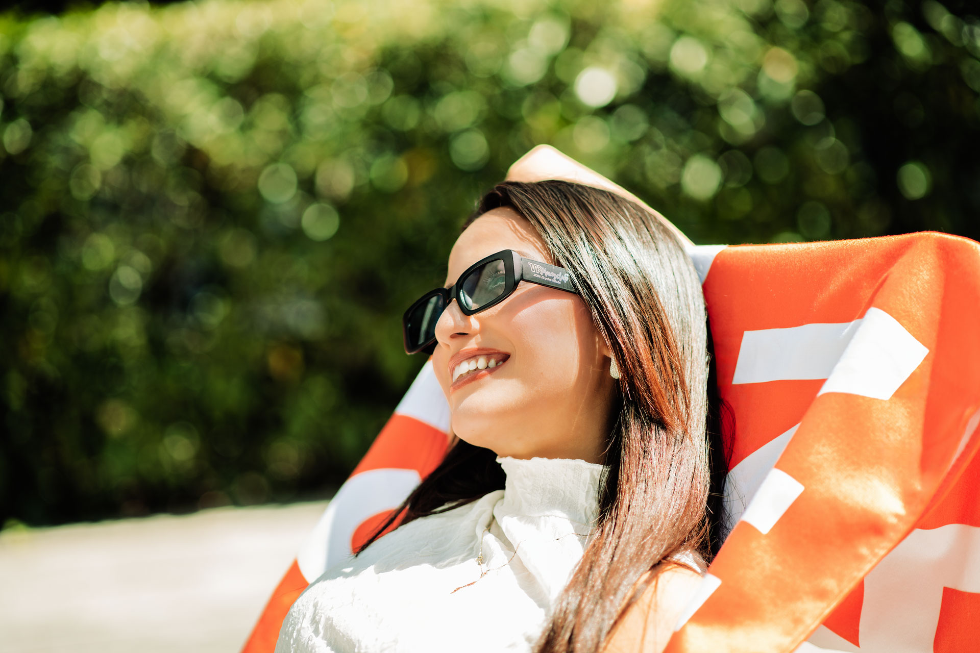 Seitenansicht einer lächelnden Person, die eine dunkle YOBA-Sonnenbrille trägt und sich entspannt gegen ein orange-weißes Handtuch mit dem VR Moneyclub-Slogan lehnt. Der Hintergrund zeigt eine sonnige, leicht unscharfe Parklandschaft.