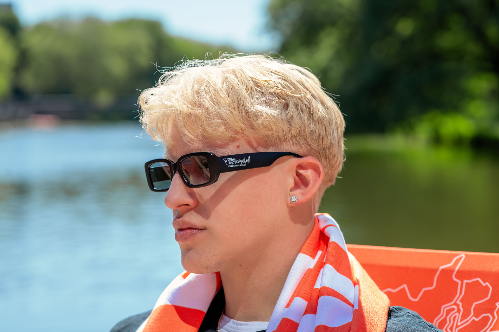 Seitenprofil einer Person mit blonden Haaren, die eine schwarze rechteckige Sonnenbrille mit weißem „VR Moneyclub“-Branding auf dem Bügel trägt. Die Person hat ein orange-weißes Handtuch um den Nacken gelegt, im Hintergrund ist ein sonniges Gewässer zu sehen.