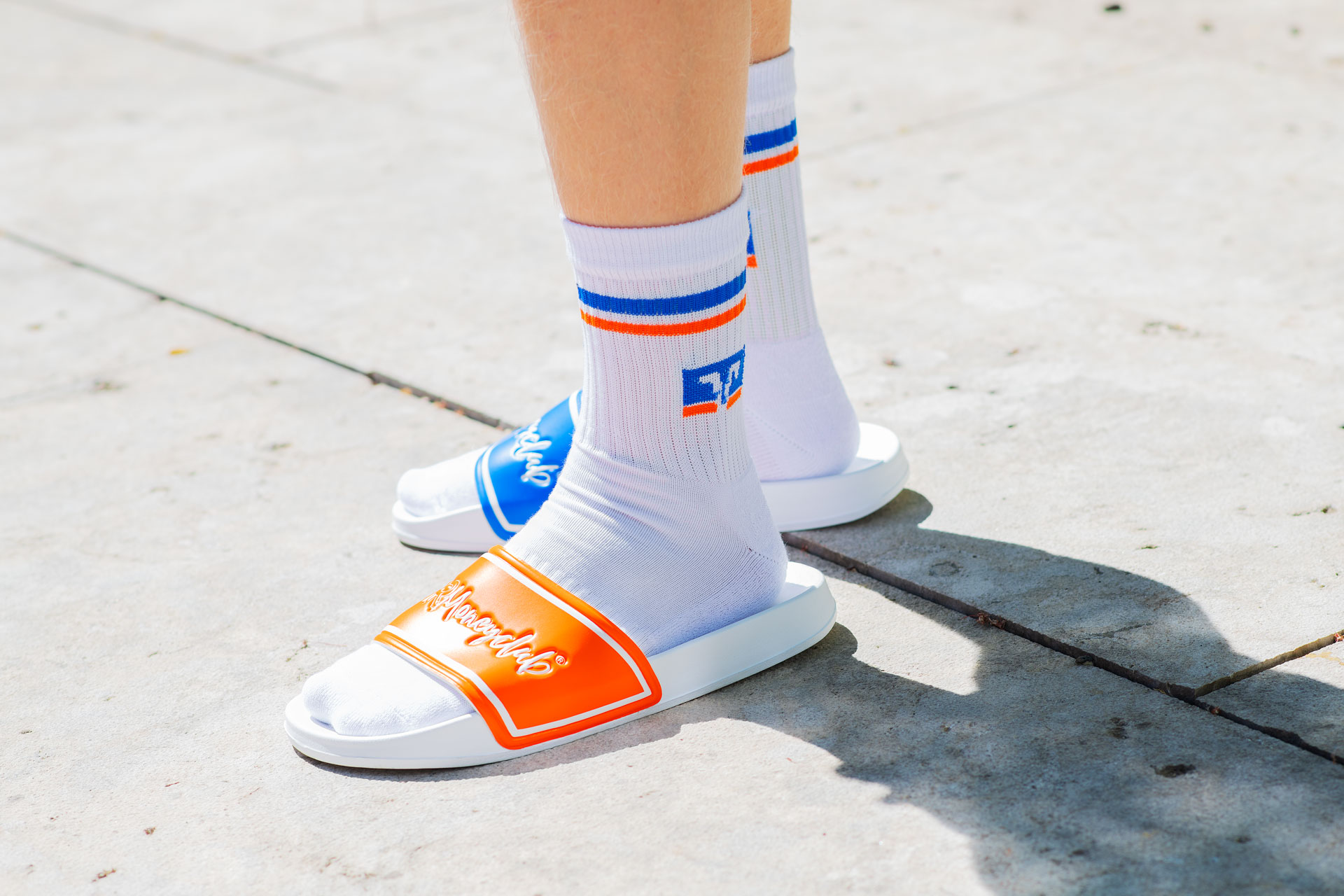 Nahaufnahme von Füßen und Unterschenkeln einer Person, die weiße Crew-Socken mit blau-orangen Streifen und VR-Logo in Kombination mit den blau-orangen VR Moneyclub Mismatch-Badeschlappen auf einem hellen Steinboden trägt.