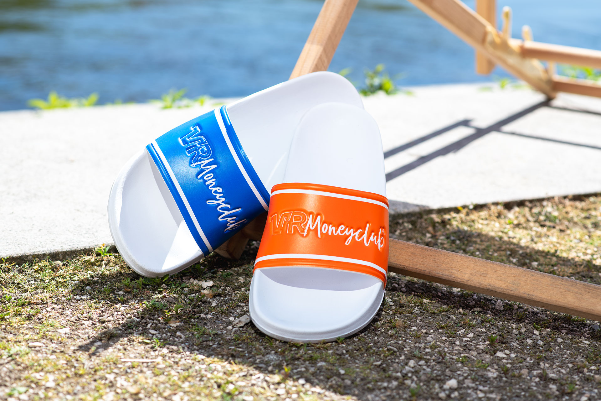 Zwei VR Moneyclub Badeschlappen in Blau und Orange im Mismatch-Design, angelehnt an einen hölzernen Liegestuhl am Wasser bei sonnigem Wetter.