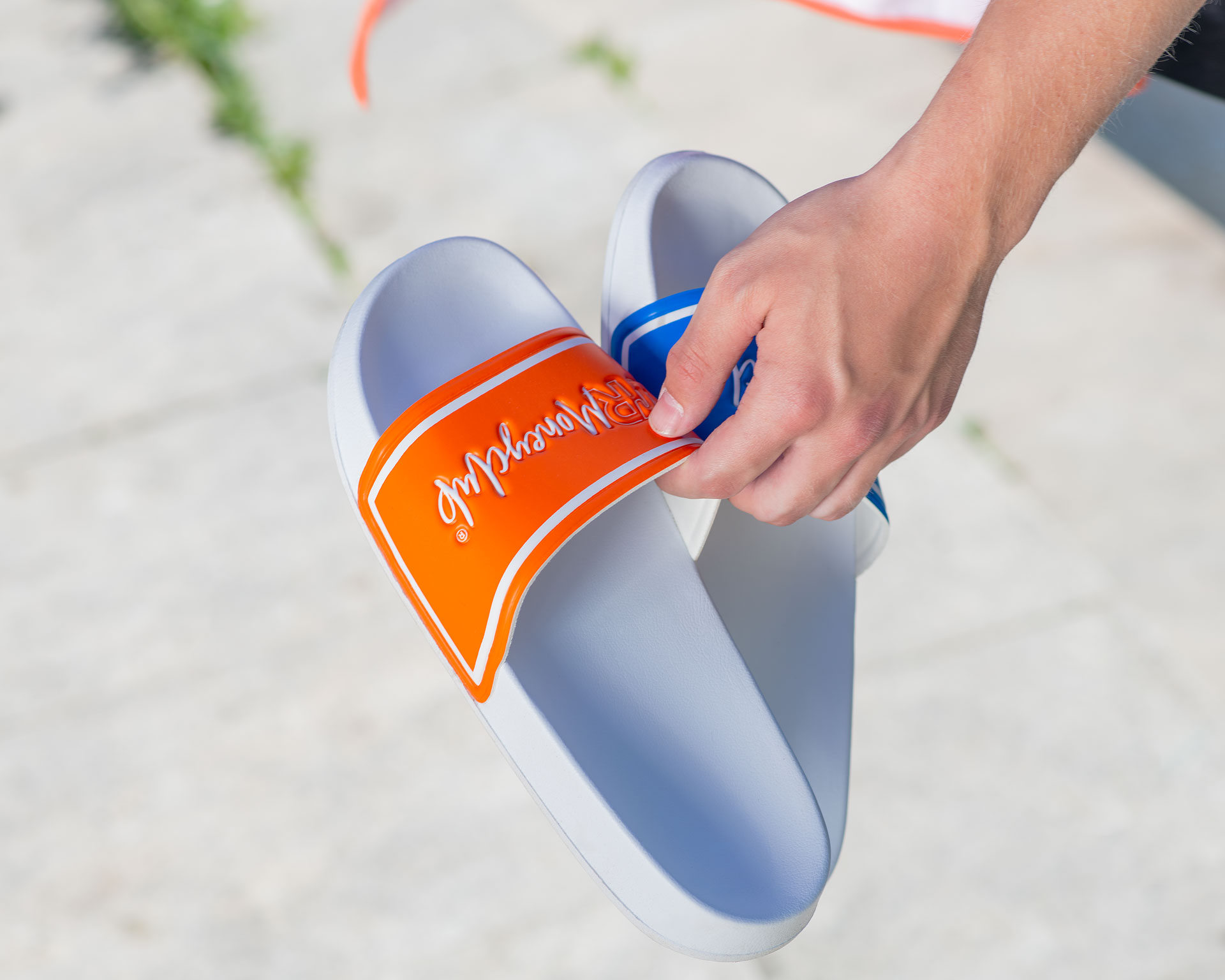 Nahaufnahme einer Hand, die ein Paar weiße VR Moneyclub Badeschlappen im Mismatch-Design (ein orangefarbener und ein blauer Riemen) hält, präsentiert in einem hellen, authentischen Lifestyle-Setting.